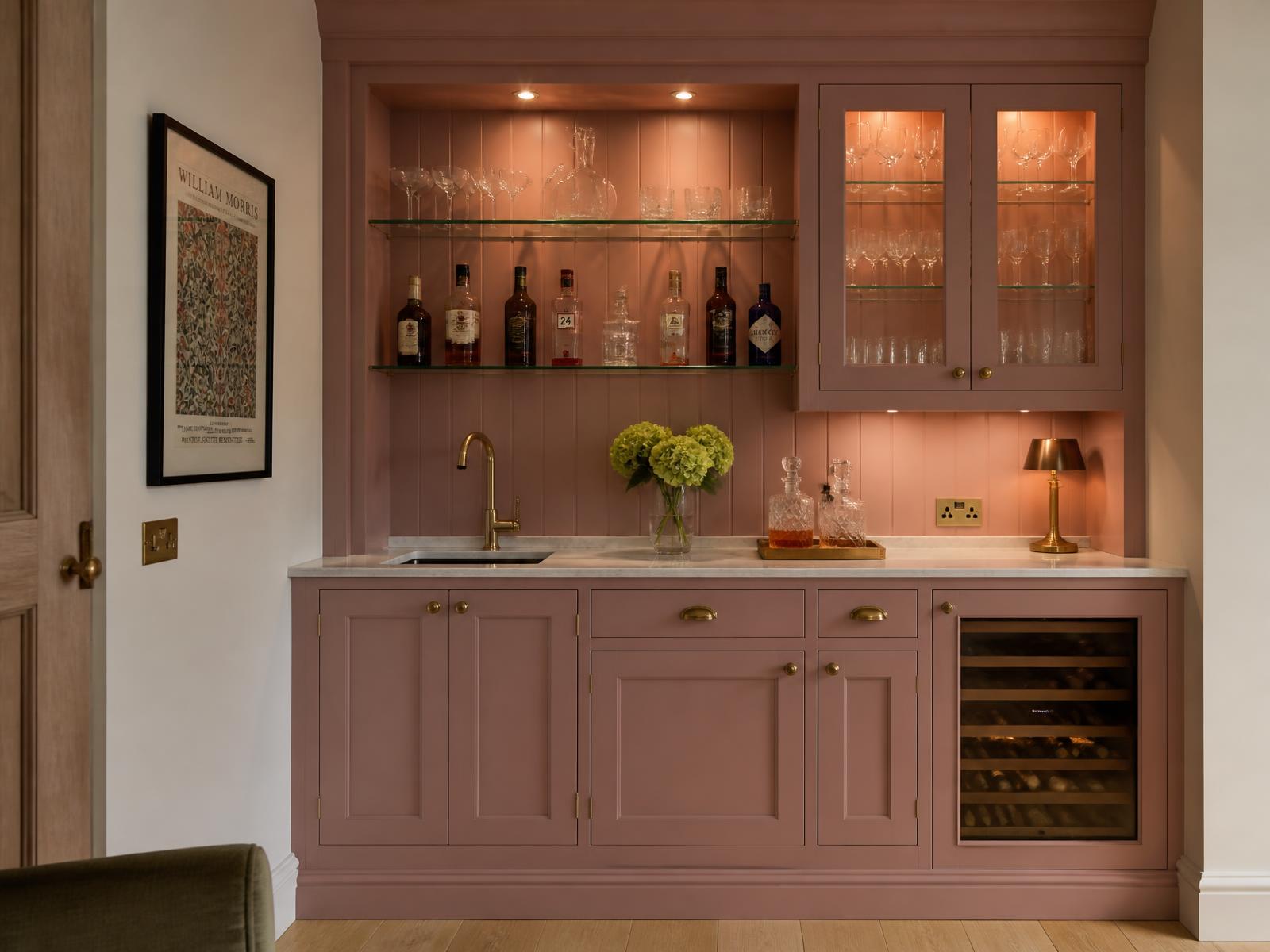 Dusty rose handmade home bar with warm lit glass shelving, brass tap, wine cabinet and marble worktop